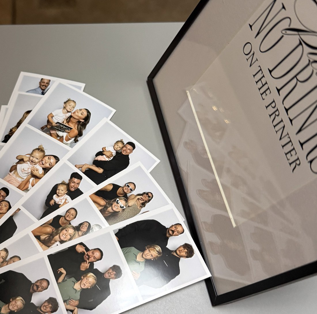 A fan of freshly printed photo strips beside a framed no drinks on the printer sign