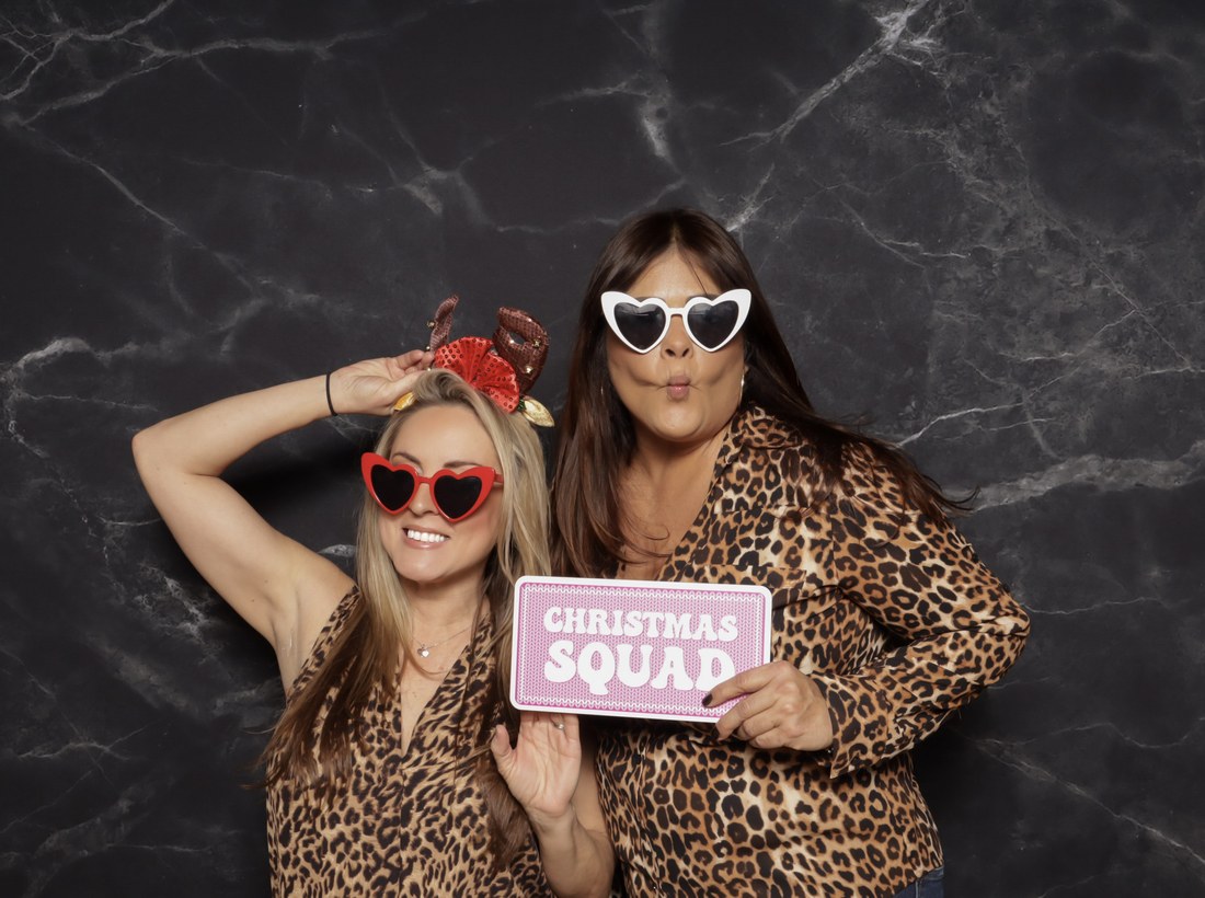 Two guests in heart shaped sunglasses holding a Christmas Squad sign in front of a black marble backdrop