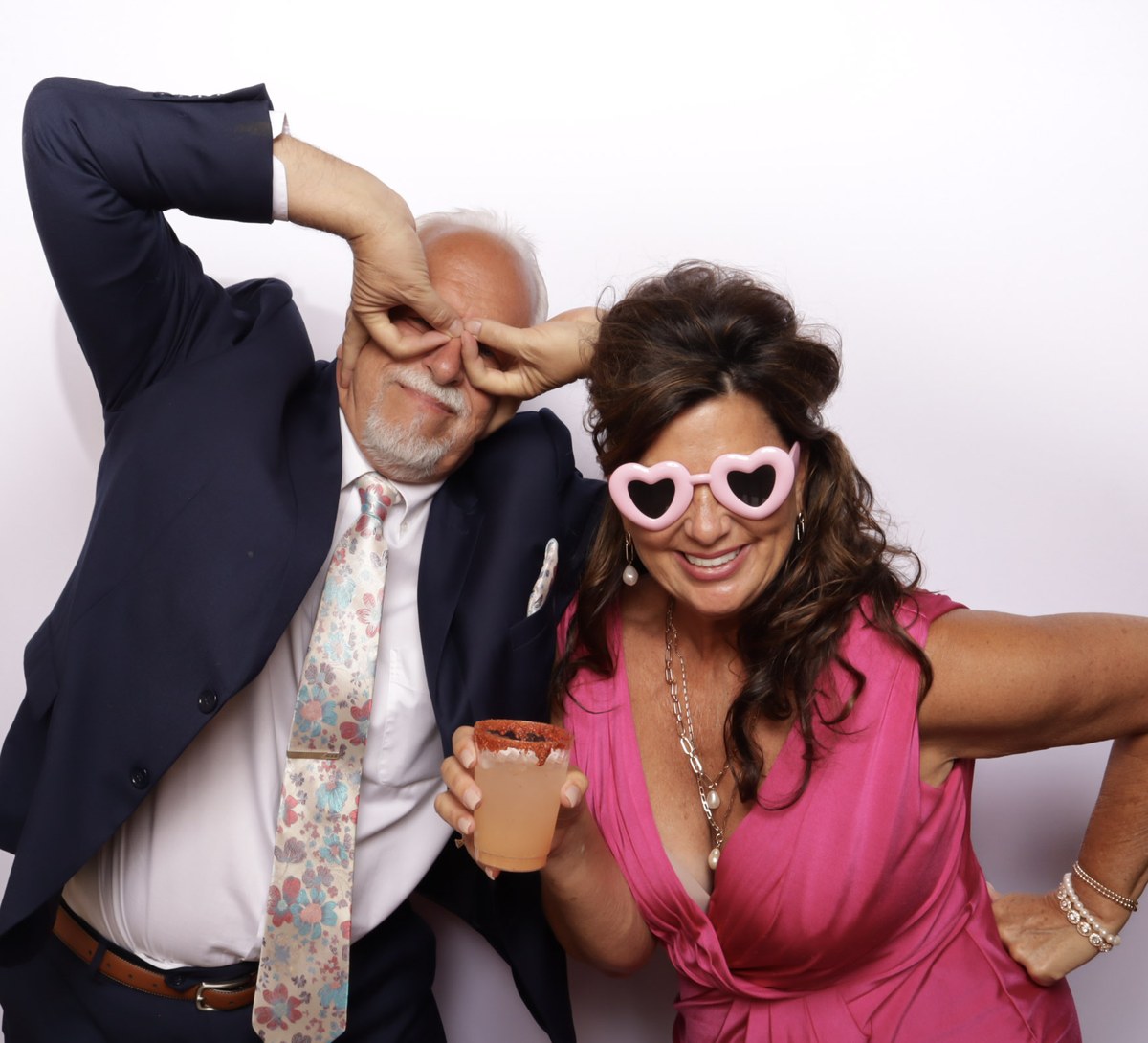 A man pulling a face beside a woman in heart shaped sunglasses holding a drink, in front of a white backdrop