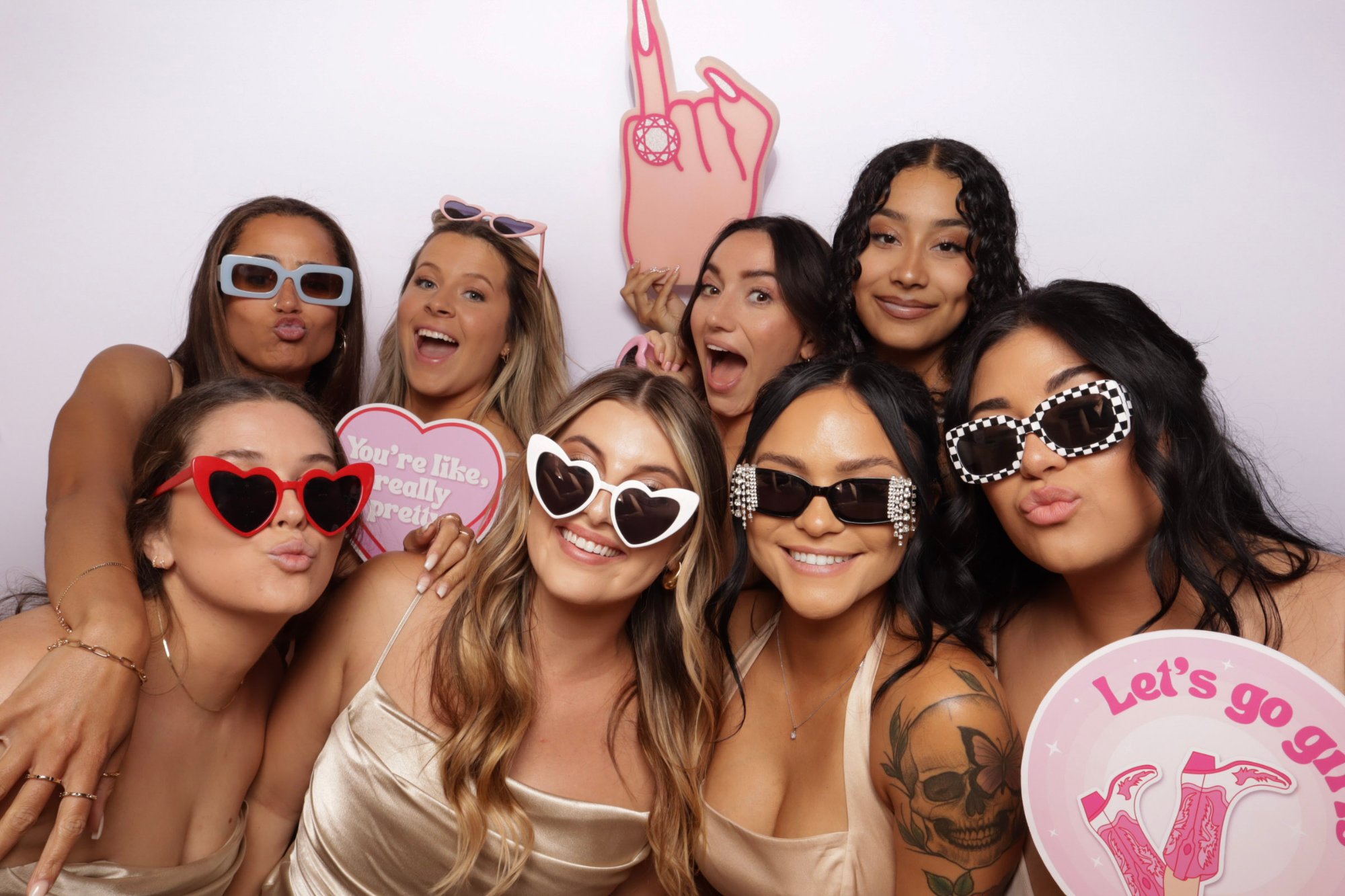 Group of women, several in satin dresses, posing with novelty sunglasses and pink party prop signs