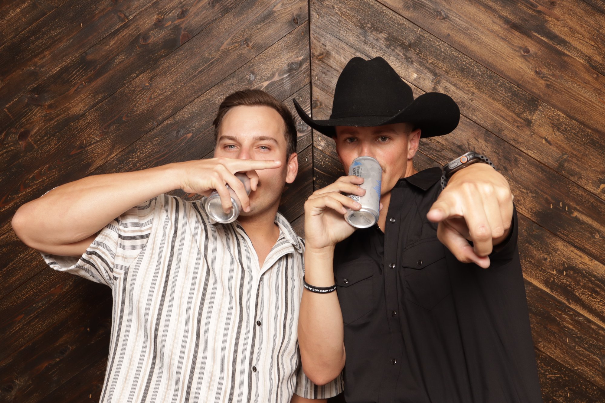 Two men sipping from cans at a dark wood backdrop, one in a cowboy hat pointing at the camera