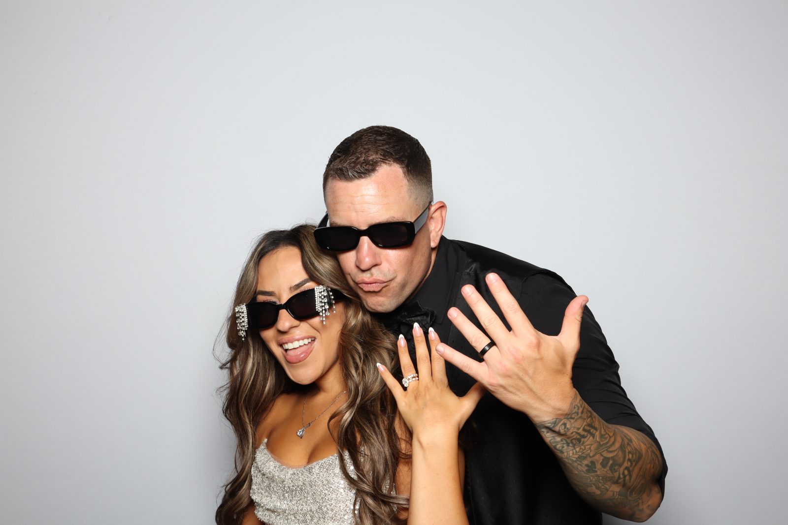 Couple in black sunglasses showing off their rings against a light grey backdrop