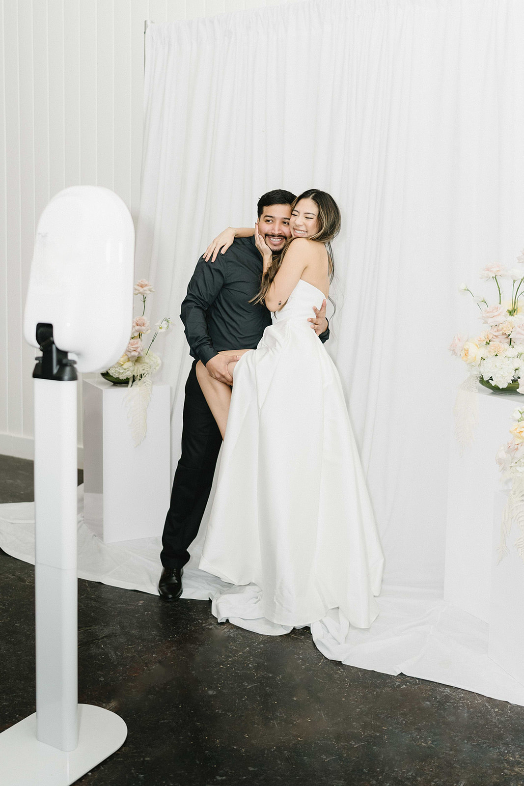 A woman in a strapless white gown hugs a man in a black shirt at a white draped backdrop beside a white photo booth