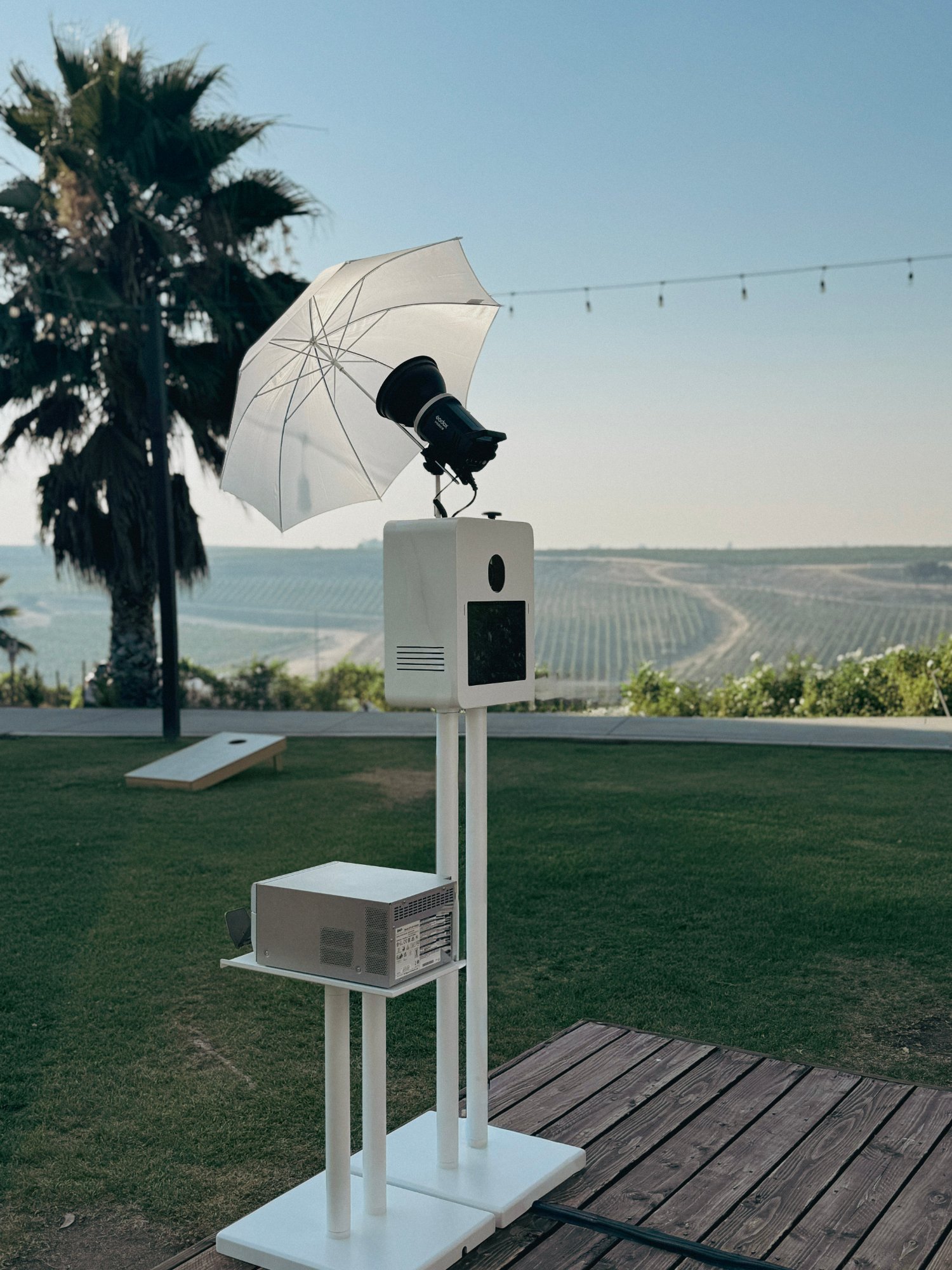 White photo booth with an umbrella light and printer on a wood deck, a lawn, palm tree and rolling farmland behind